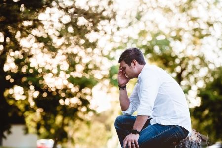 A man in a white button down shirt and jeans sits on a stump in the woods, leaning on his knee with one hand and with his head in the other hand.